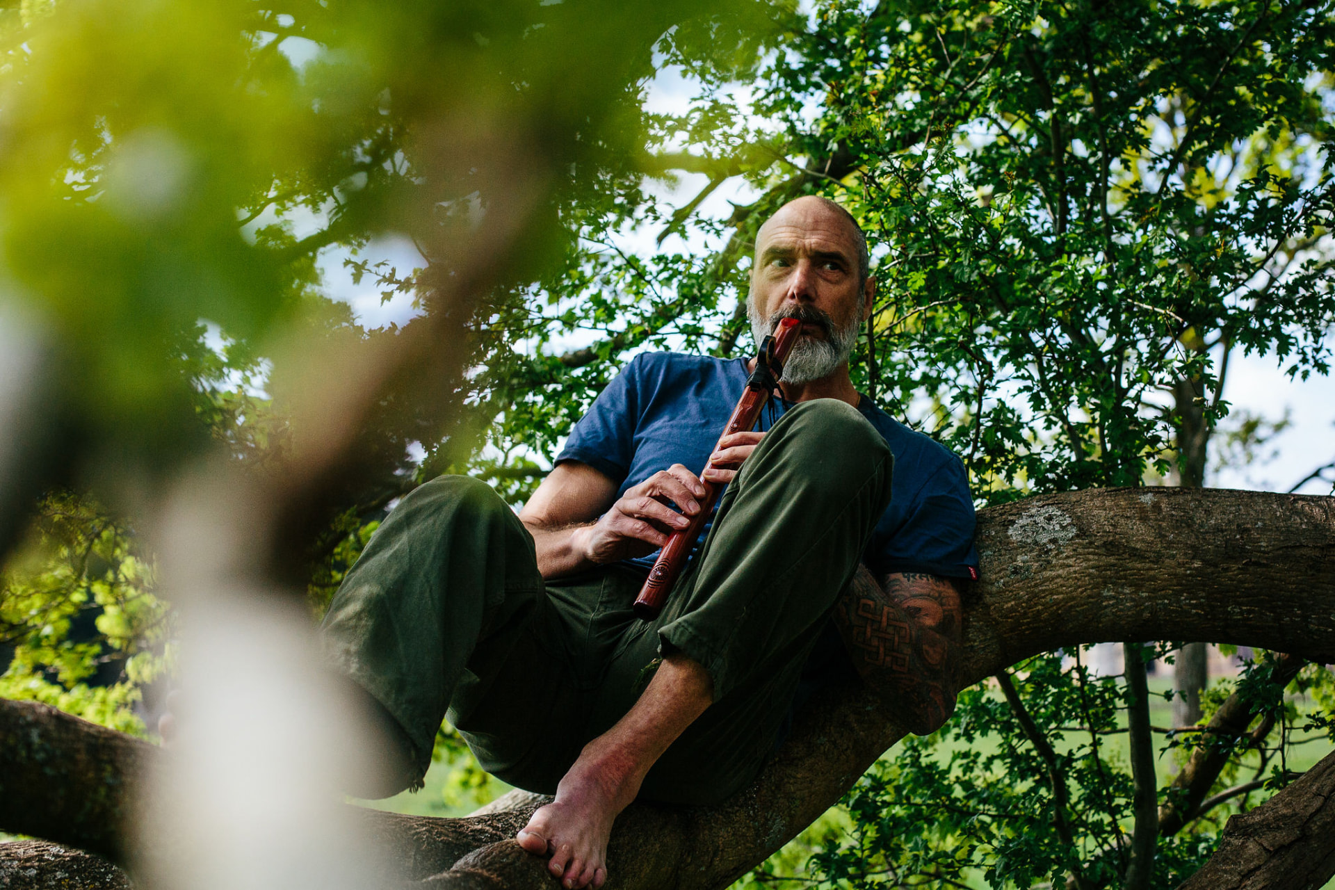 Christopher Gladwell seated in a tree playing a wooden flute
