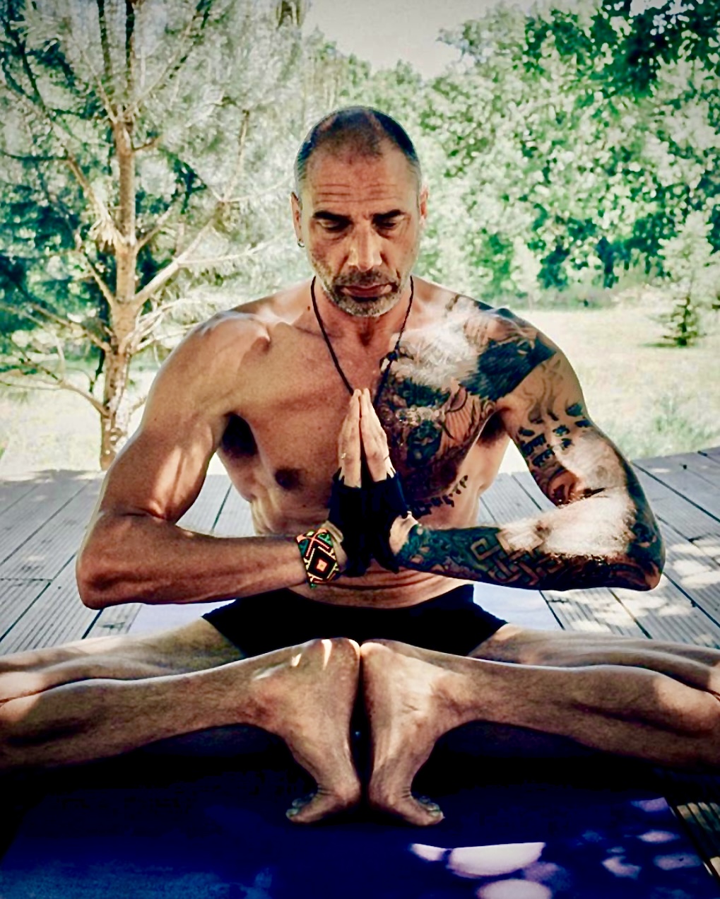 Christopher Gladwell seated in a wide-legged yoga posture with hands in prayer mudra on an outdoor deck
